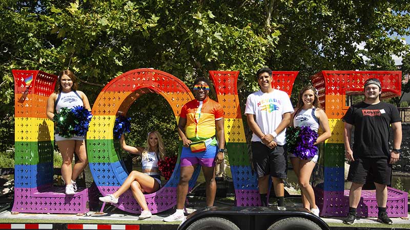 A group of people, including two Nevada cheerleaders and some wearing University of Nevada, shirts, stand inside of and next to giant letters "LOVE" that are painted with the LGBTQ+ rainbow of colors, red, yellow, purple, green and indigo.